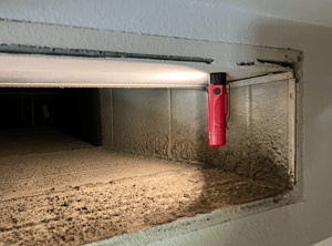 A red flashlight shines line into a duct that is heavily dusty.