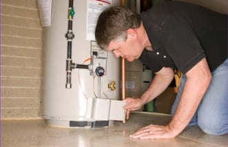 A man is crouched down with his left hand flat on the floor and his right hand holding an access panel of a conventional water heater.