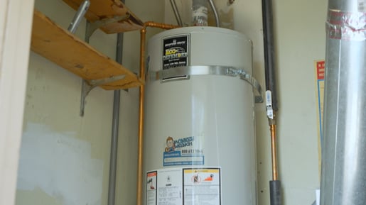 A tank-style water heater installed in a home with a Monkey Wrench Plumbing, Heating & Air sticker. 