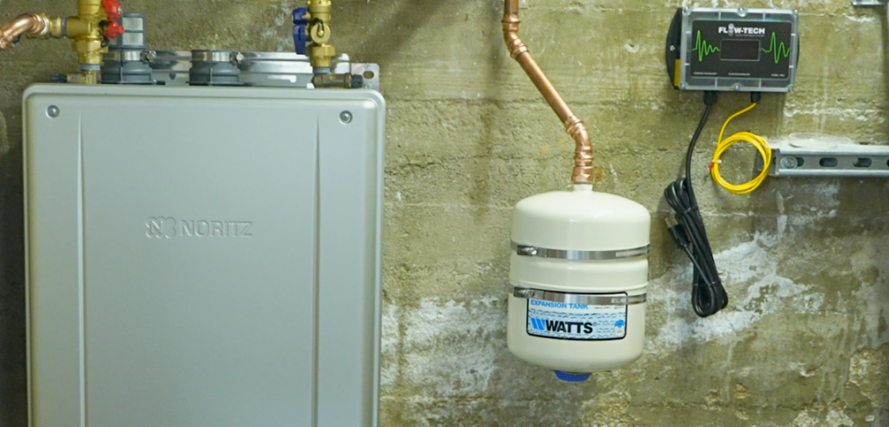 A Noritz tankless water heater with a expansion tank to the right and a flowtech device in the top right corner.