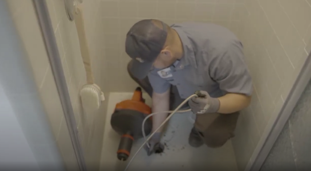 The image shows a Monkey Wrench technician clearing a shower drain with a drain snake.