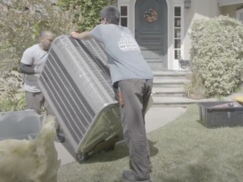 The image shows two Monkey Wrench technicians lifting an HVAC unit onto a dolly.